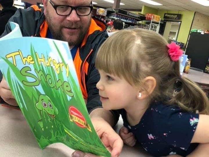A father reading The Hungry Snake to his young daughter at a table, the pair of them looking at the open book.