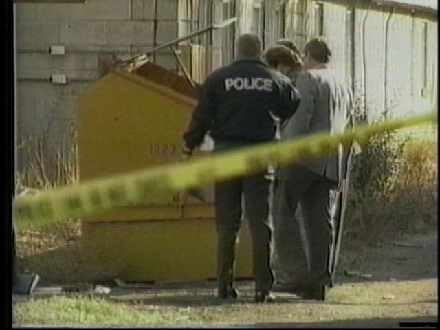 A police officer and two investigators standing beside a dumpster behind a building, with crime scene tape strung across the foreground.