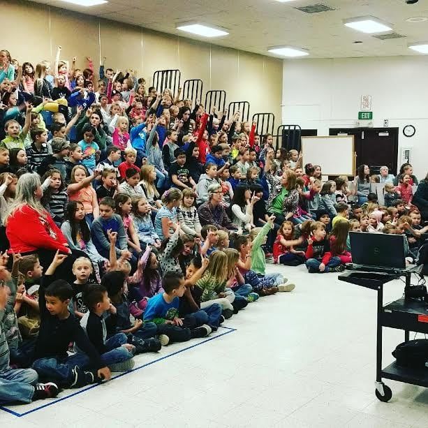 Suzanne Hobbs speaking to a full school assembly of children seated on the floor.